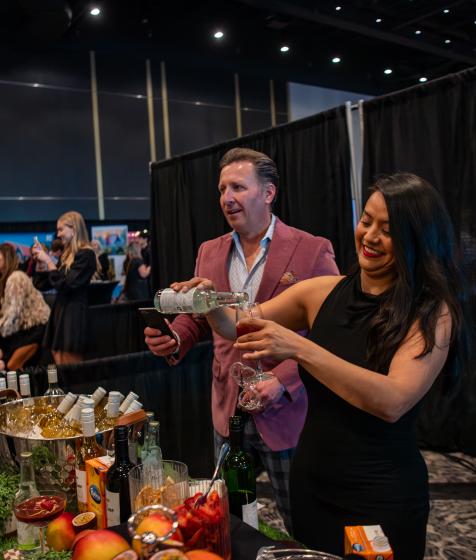 A female mixologist with dark hair and a black dress pours a drink from a martini shaker into a glass in front of a large crowd at the Convention Centre