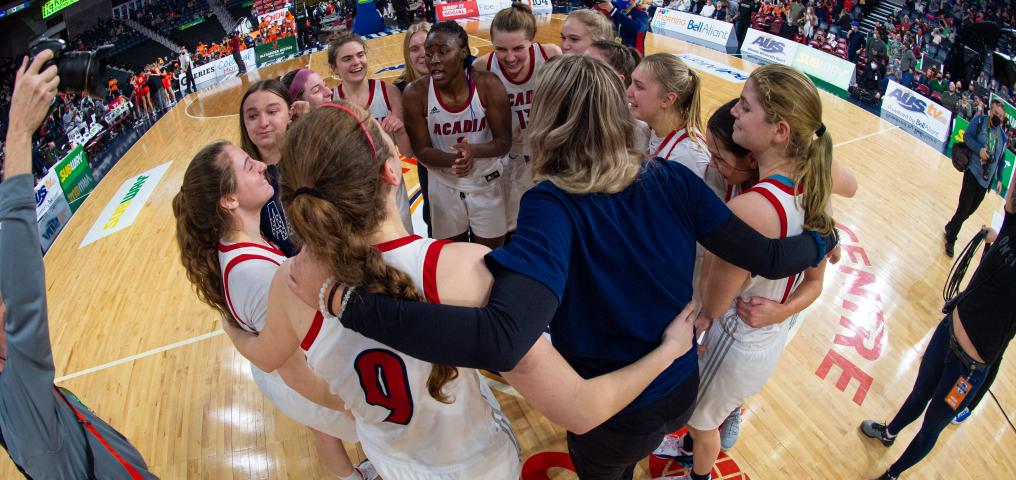 a 2022 women's team is shown huddling off the court side with the coach's arms holding them together